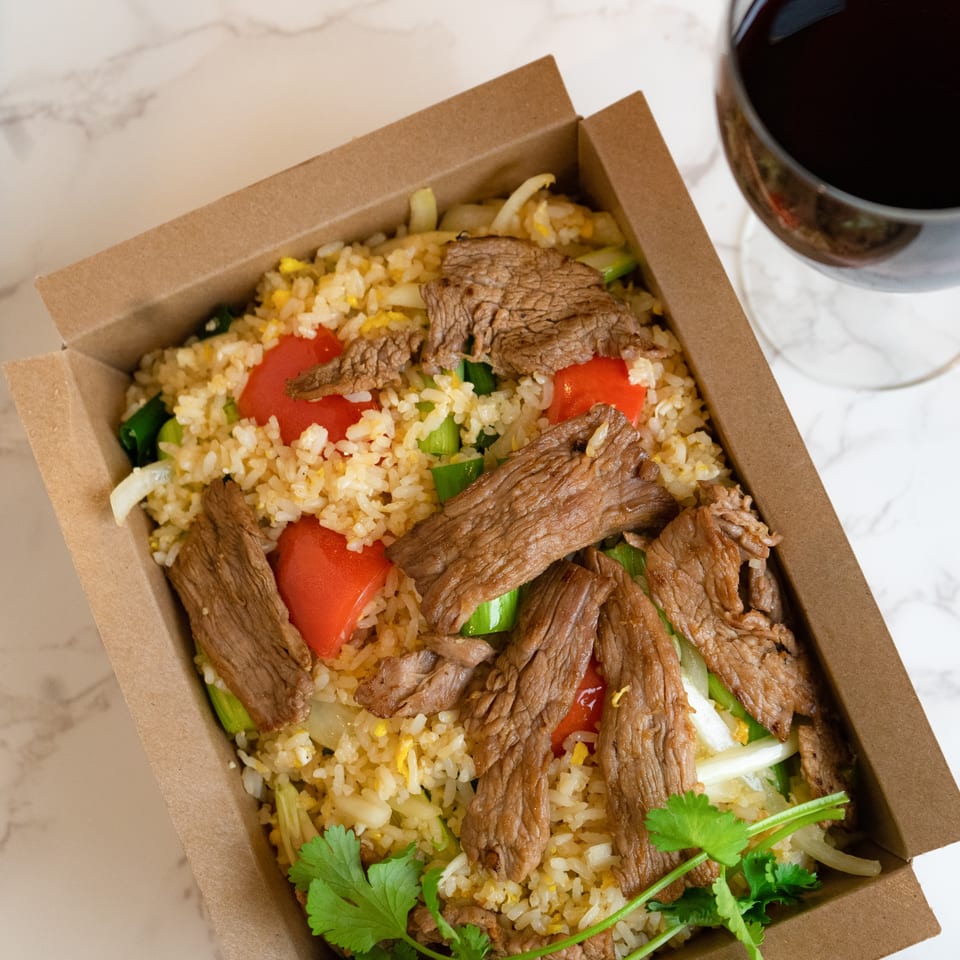 Thai beef fried rice with red peppers and cilantro in a takeout box, next to a glass of red wine.