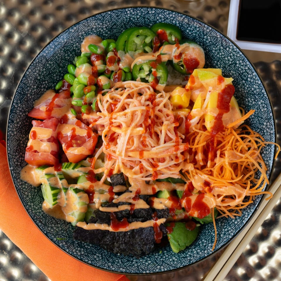 Colorful Thai poke bowl with edamame, jalapenos, mango, carrots, tomatoes, and noodles drizzled with sauce.