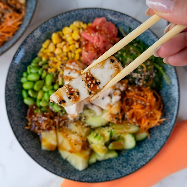 Chopsticks lift a piece of chicken from a colorful Thai bowl with corn, edamame, and carrots.