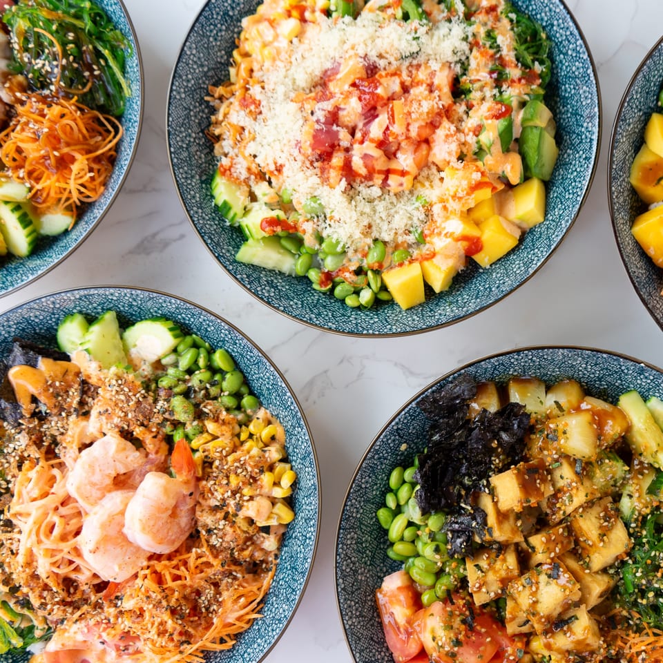 Overhead view of five colorful Thai poke bowls with various toppings on a white marble surface.