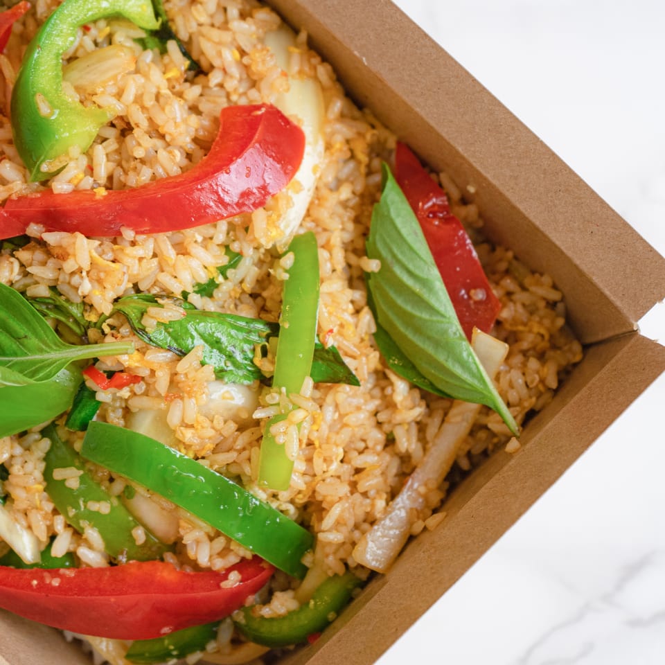 Close-up of Thai fried rice with red and green bell peppers, basil, and onions in a takeout box.