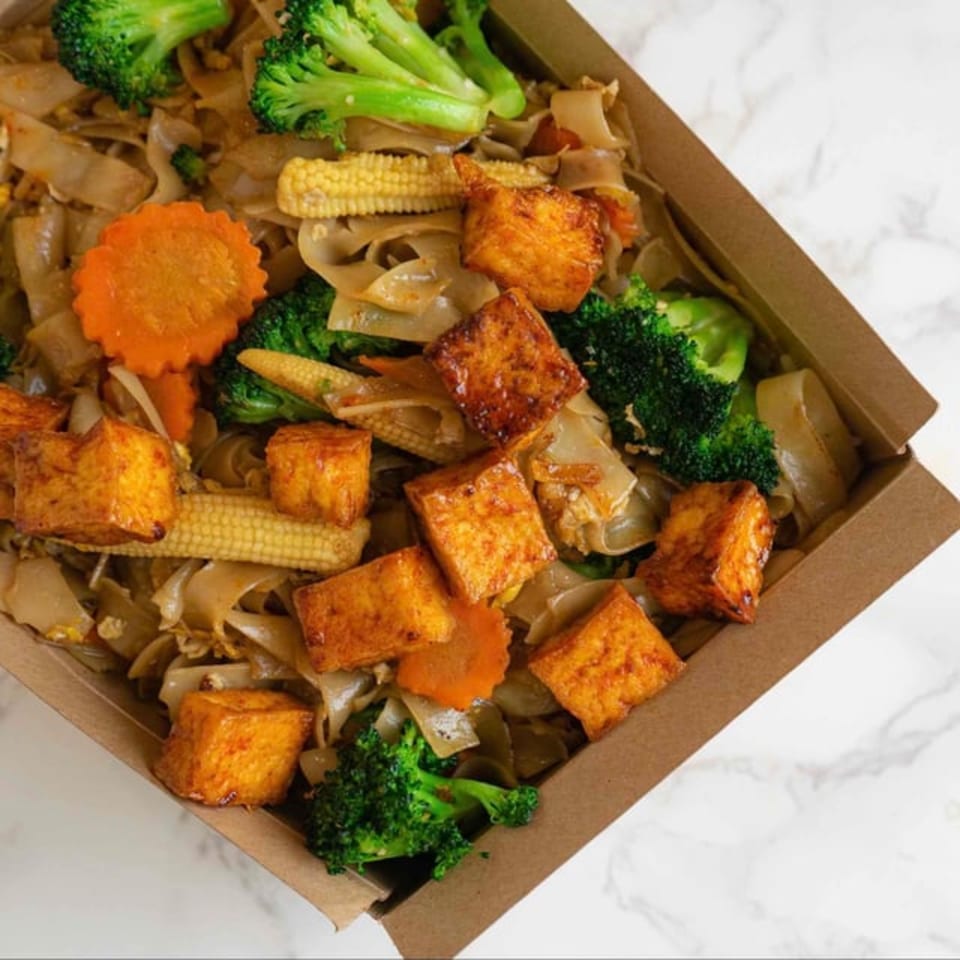 Thai noodles with crispy tofu, broccoli, baby corn, and carrots in a takeout container.