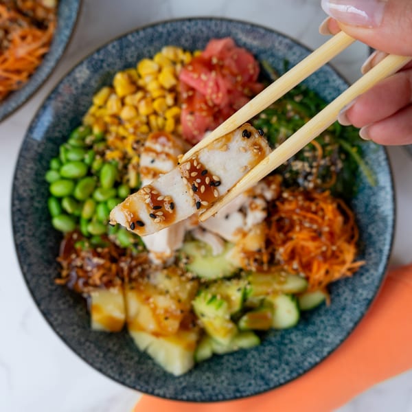 Chopsticks lifting a piece of chicken from a colorful Thai bowl with corn, edamame, and carrots.