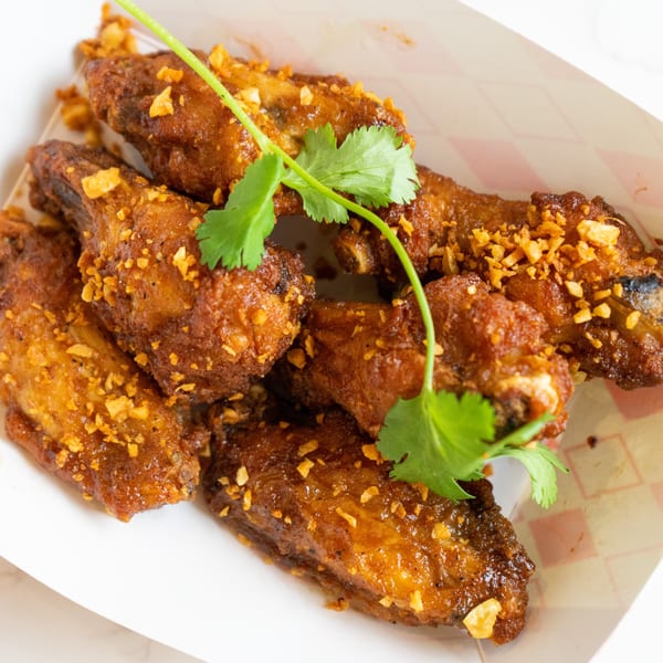 Crispy Thai chicken wings, garnished with cilantro and fried garlic, served in a paper tray.