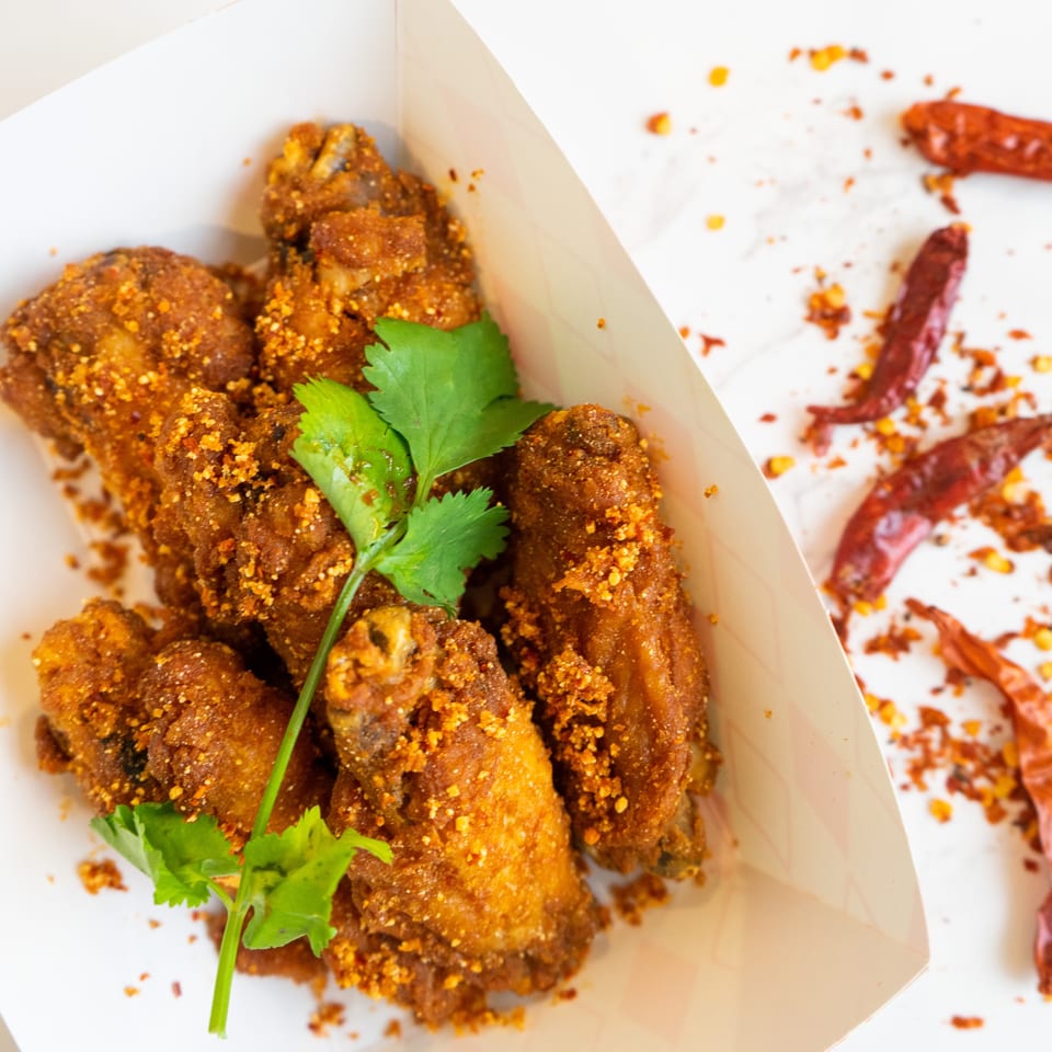 Crispy Thai fried chicken wings with chili flakes and cilantro in a white paper tray.